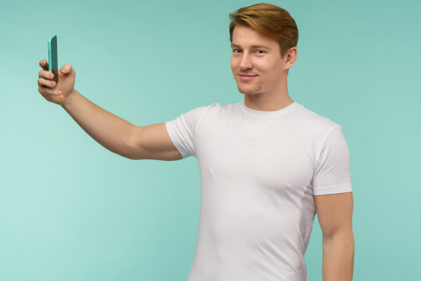 帅气的红发运动男在用蓝色背景的智能手机自拍或在线广播。图片下载