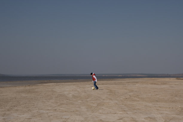 一个穿着白色运动衫和橙色背心的10岁男孩在荒芜的海滩上孤独地踢足球。图片下载