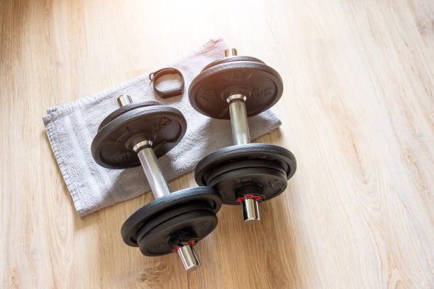 灰色毛巾上放着两个哑铃和一个带时钟的健身手镯，在家锻炼。图片下载