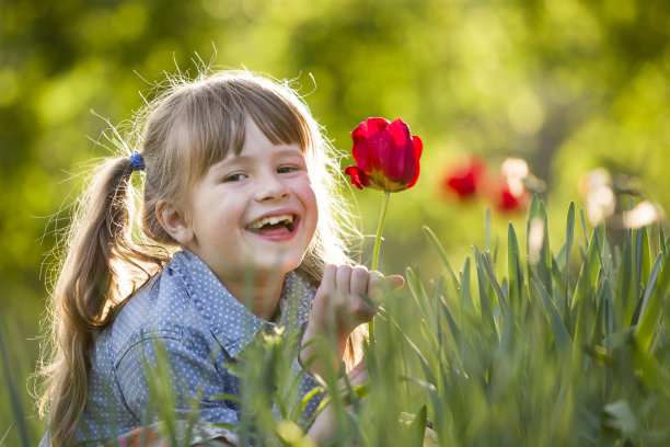 可爱的漂亮微笑的孩子女孩与灰色的眼睛和长头发与明亮的红色郁金香花在模糊阳光下的绿色散焦背景。热爱自然的理念。图片下载