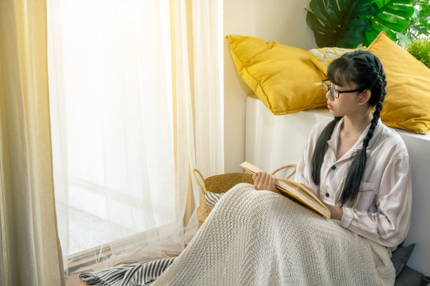 年轻美丽的亚洲少女穿着睡衣和眼镜独自坐在卧室地板上享受读书。放松女生与家庭学校学习期间的社会距离隔离。图片下载
