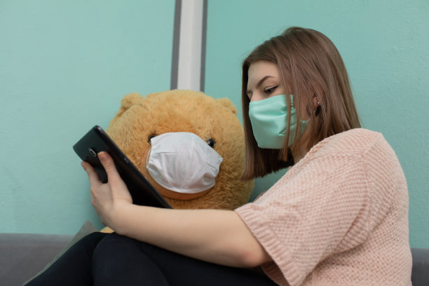 这个小女孩和大泰迪熊一起为考试学习。他们都戴着防护口罩。图片下载