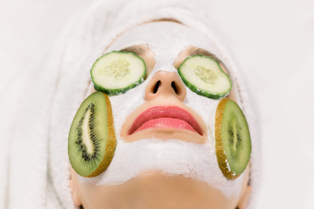 近距离微距照片的年轻女子与自制面膜从天然成分保湿皮肤，新鲜黄瓜片在她的眼睛，猕猴桃片在脸颊，和头发包裹在浴巾图片下载