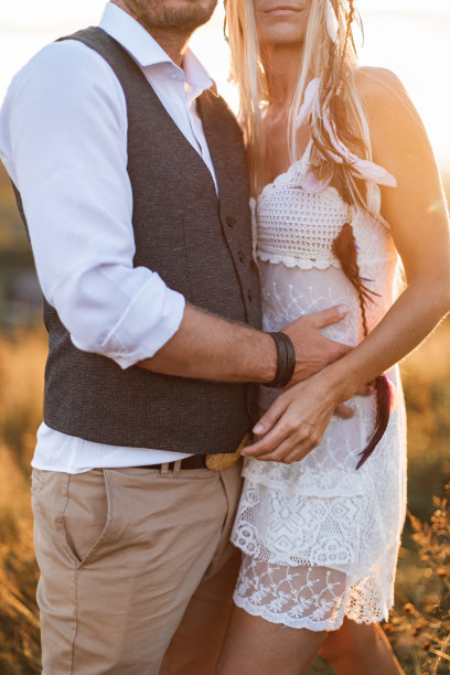 裁剪的照片美丽的夫妇穿着波西米亚风格拥抱对方在阳光明媚的夏日田野。英俊的男人穿着休闲装，而女人穿着白色的连衣裙和羽毛的头发在田野里行走图片下载