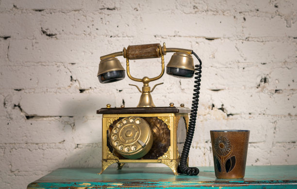 绿色的老式木桌和白色的砖墙上放着金色的古董电话机和陶瓷马克杯图片下载