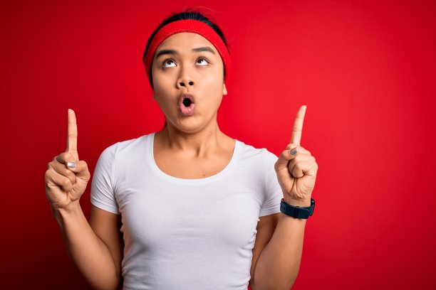 年轻美丽的亚洲运动女人穿着运动服做运动在红色的背景惊讶和惊讶向上看和指向手指和举起的手臂。图片下载