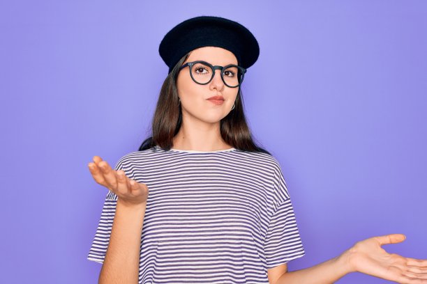 年轻美丽的黑发女人戴眼镜和法国贝雷帽在紫色的背景愚蠢和困惑的表情与胳膊和手举起。怀疑的概念。图片下载