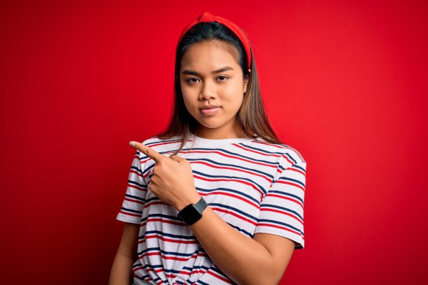 年轻美丽的亚洲女孩穿着休闲条纹t恤，孤立的红色背景指向与手指的一边显示广告，严肃而冷静的脸图片下载