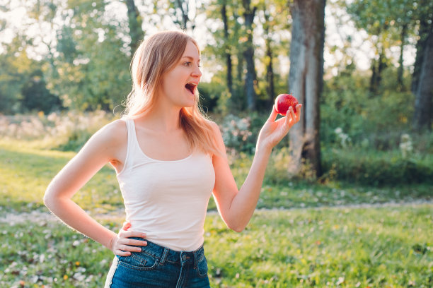 迷人的年轻女子在模糊的自然背景上吃红苹果。在公园吃午饭。人类情感的概念。有趣的饥肠辘辘的女孩图片下载