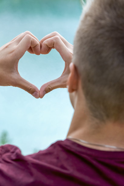 一个年轻人的特写，使一个心的形状与他的手与绿色的水的海滩。图片下载