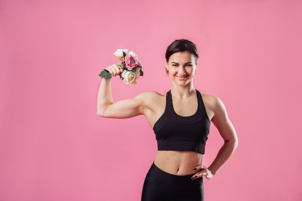 这是一张有趣的照片，一个年轻的肌肉健康的女孩手里拿着一束花，闻着花香，锻炼肌肉。快乐微笑的欧洲运动女人，展示二头肌在粉红色的背景图片下载