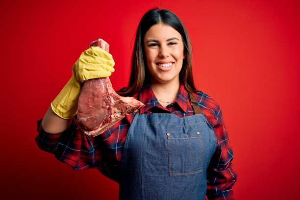 年轻的屠夫女人拿着新鲜的生牛肉桩子在红色的背景与一个快乐的脸站着，微笑着自信的微笑显示牙齿图片下载