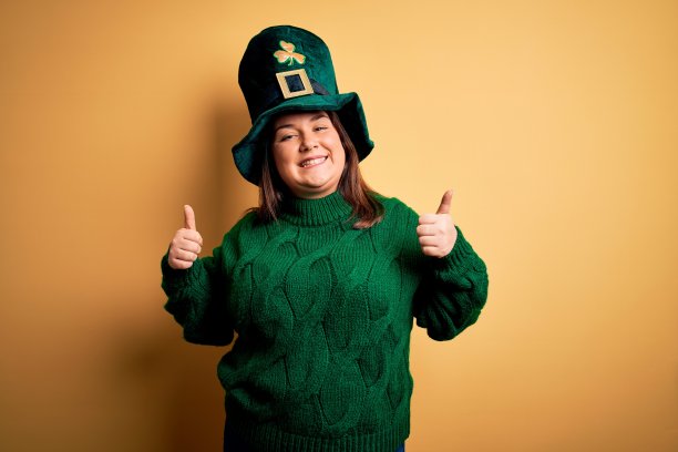 年轻美丽的大尺寸女人戴着绿帽子与三叶草庆祝圣帕特里克节成功手势，拇指向上微笑和幸福。愉快的表情和胜利者的姿态。图片下载