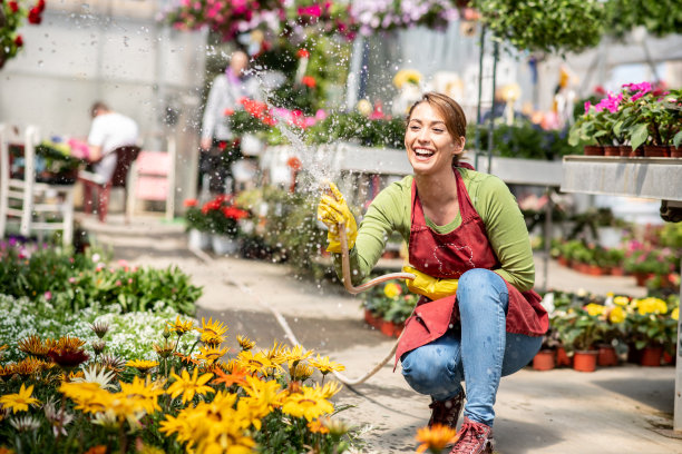 快乐的女园丁浇盆花与花园软管在苗圃。图片下载
