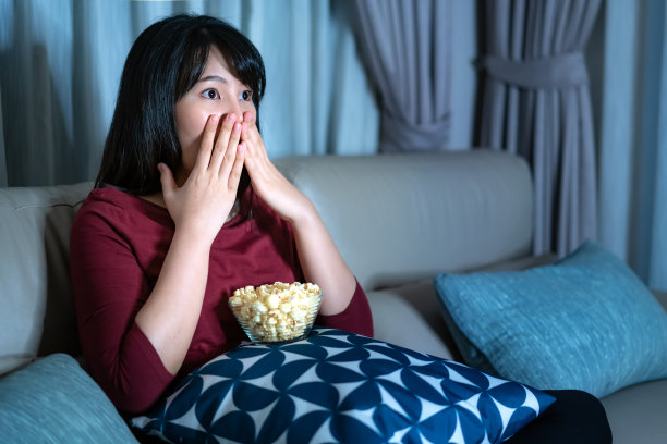 年轻的亚洲女人看电视悬疑电影或新闻，看起来震惊和兴奋深夜在家里客厅的沙发上吃爆米花。图片下载