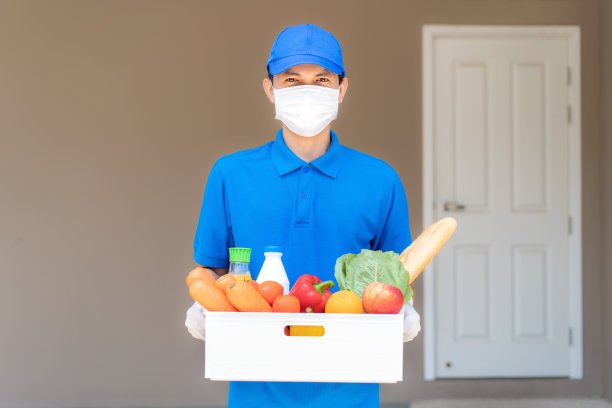 在居家隔离期间，一名亚洲快递员戴着口罩和手套，手拿装有食品、水果、蔬菜和饮料的杂货箱，站在顾客家门口。图片下载