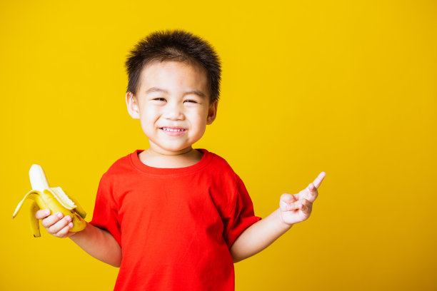 快乐肖像亚洲小孩或小孩可爱的小男孩迷人的微笑穿着红色的t恤玩着剥了皮的香蕉吃，工作室拍摄孤立在黄色的背景图片下载
