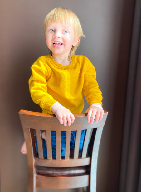 一个皮肤白皙的孩子坐在一把木椅上，在一堵棕色的墙壁上摆姿势。图片下载