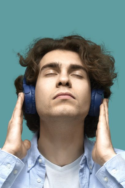 一个年轻漂亮的男子的肖像穿着白色t恤和蓝色衬衫在蓝色耳机。他高兴地闭上了眼睛图片下载