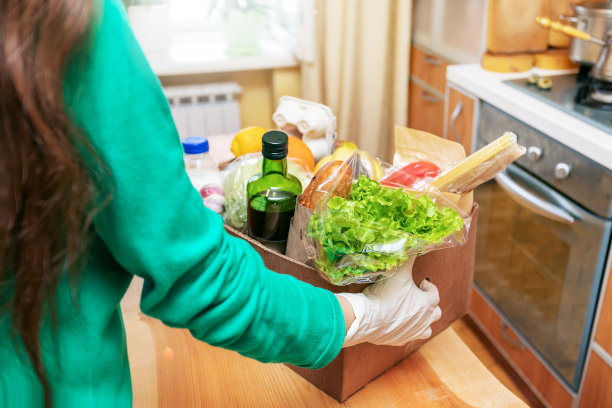 穿着绿色帽衫和防护手套的妇女把装有食品的盒子放在厨房的木桌上。有选择性的重点。图片下载