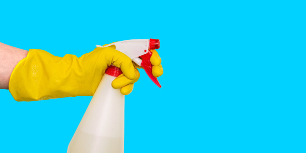 在一个黄色的手套拿一个喷雾器。蓝色背景上的瓶子。喷雾器用于办公室和家庭的清洁和消毒。侧视图，拷贝空间。图片下载
