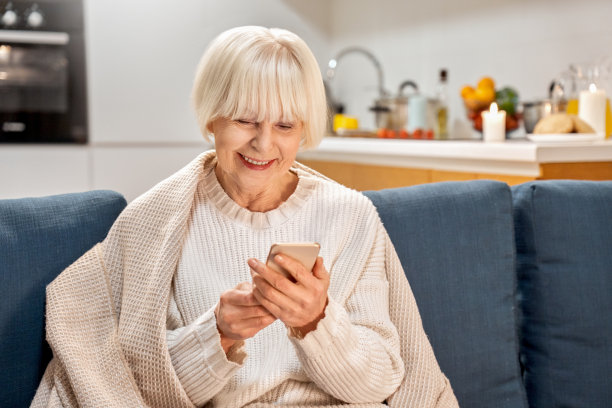 一位穿白色衣服的资深女士坐在沙发上。友善有趣的老奶奶等着孩子，网上聊天上网笑开心的女士老。成人独女在手机上看，使用数字app。图片下载