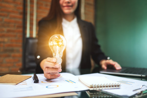 一位女商人在办公室里用笔记本电脑工作，手里拿着一个发光的灯泡图片下载