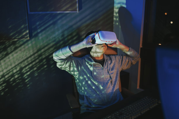 一名亚裔中国男性，晚上在他的家庭办公室自习室，戴上VR眼镜，在他的台式电脑前体验3D虚拟游戏图片下载