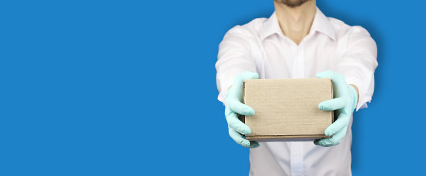 手拿医用手套举着纸板箱，概念邮件速递货物在病毒检疫，横幅有拷贝空间，蓝色背景图片下载