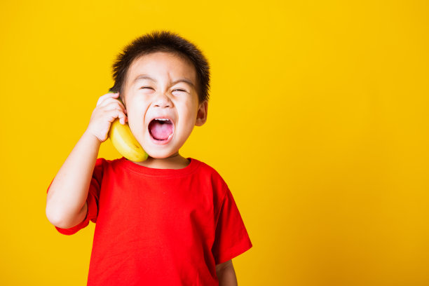 快乐肖像亚洲小孩或小孩可爱的小男孩迷人的微笑穿着红色的t恤玩拿着香蕉水果假装像电话，工作室拍摄孤立在黄色的背景图片下载