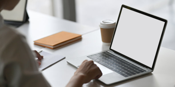 裁剪的图像美丽的男子使用一台电脑笔记本与白色空白屏幕，而坐在白色的办公桌在舒适的客厅作为背景。图片下载