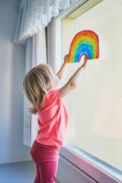 可爱的蹒跚学步的小女孩把彩虹的图画贴在窗户玻璃上图片下载