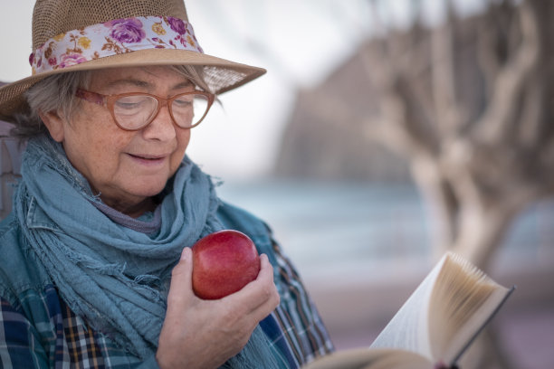 老年妇女享受退休生活的肖像，坐在海滩边看书，吃苹果。水面上的地平线。海洋景观。一个只有戴眼镜和帽子的人图片下载