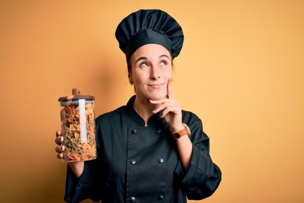 年轻漂亮的女厨师穿着炊具制服，戴着帽子拿着意大利面瓶子严肃地面对思考问题，非常困惑的想法图片下载