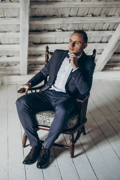 时尚的新郎肖像背景的白色木墙。乡村婚礼理念在谷仓，文字空间。波西米亚新婚快乐。男人在老式椅子上放松图片下载