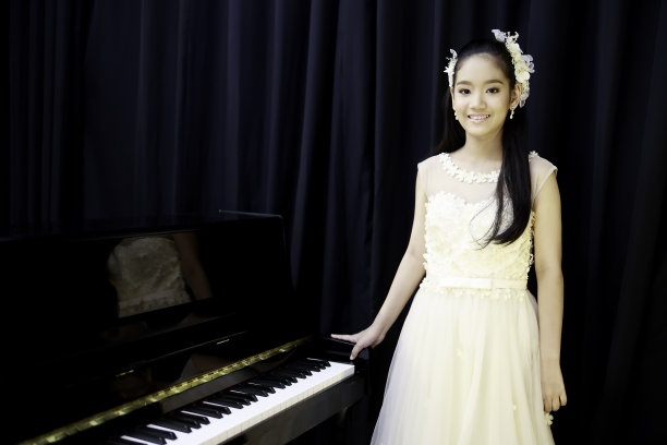 一个亚洲女孩化了妆，梳了发，穿着白色的晚礼服为钢琴表演。图片下载