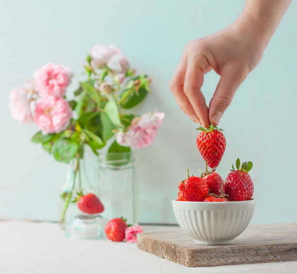 一位女士把一颗草莓放在装满鲜红多汁的草莓的水果盘里。色彩柔和的装饰，以薄荷色为背景，点缀花卉。图片下载