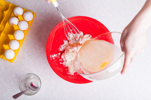 女士一手拿着打蛋器，另一只手把打好的鸡蛋倒入透明的玻璃碗里，在红色的盘子里准备面团做馅饼，旁边的桌子上是一杯稀释的苏打水。图片下载