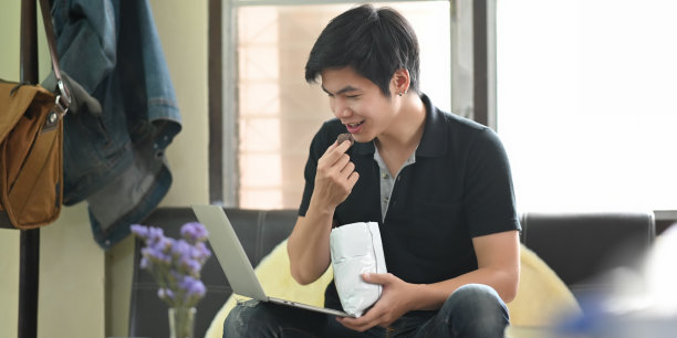 这张照片是一个聪明的男人手里拿着零食，看着笔记本电脑，坐在舒适的客厅的皮沙发上作为背景。一名男子在家中休养生息。图片下载