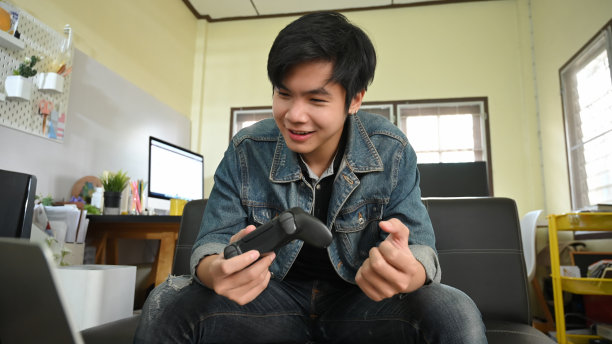 这是一个聪明的男人坐在舒适的客厅的皮沙发上玩电子游戏，手里拿着操纵杆的照片。一名男子在家中休养。图片下载