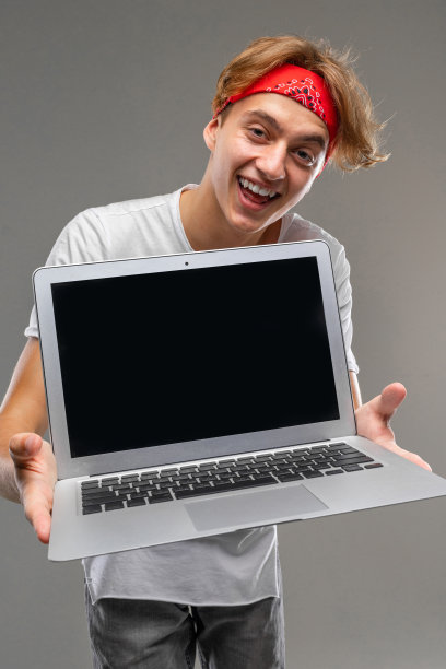 一个戴着大手帕、带着笔记本电脑的快乐男人的垂直镜头，空白屏幕前，在灰色孤立的背景上，他咧着嘴笑着，露出牙齿图片下载