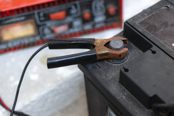 一个汽车电池跳线与黑色减铜钳连接到汽车电池的特写。电池充电器在后台。图片下载
