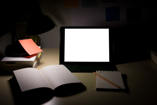 写字台上放着空白屏幕的写字板，旁边放着书本和文具。晚上在家办公。COVID-19大流行期间的在线学习和在家工作概念。图片下载