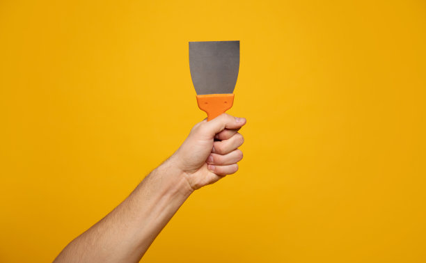 刮腻子。特写照片的手拿着一个腻子刮刀在一个黄色的背景。家庭装修的概念。图片下载