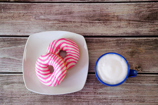 木桌上放着一个盘子，里面有两个粉色的甜甜圈和一个蓝色的杯子，里面有新鲜的卡布奇诺。图片下载