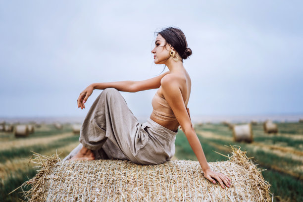 在温暖的秋日里，赤着脚，浅黑色的亚麻裤子和裸露的肩膀坐在干草堆上。她身后是一片麦田图片下载