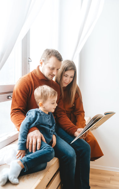 微笑的父母和带书的小男孩在家里的窗户附近。图片下载