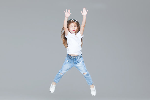 学龄前的小女孩穿着蓝色牛仔裤，白色t恤和太阳镜，举起的手孤立在灰色背景上跳跃图片下载