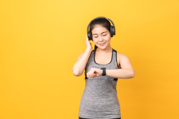 微笑的亚洲女人穿着运动装看起来智能手表和耳机在橙色的背景。图片下载