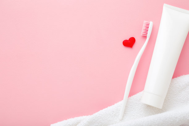 牙刷，白色牙膏和毛巾放在浅粉色的桌子上。柔和的颜色。亮红色的心。爱健康牙齿的理念。文字或标识的空白位置。特写镜头。自顶向下的观点。图片下载